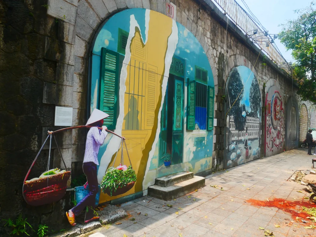 Vietnam 62 Mural vibrante en Vietnam que muestra una figura con un sombrero tradicional y cestas de verduras, rodeado de coloridos murales en la pared.